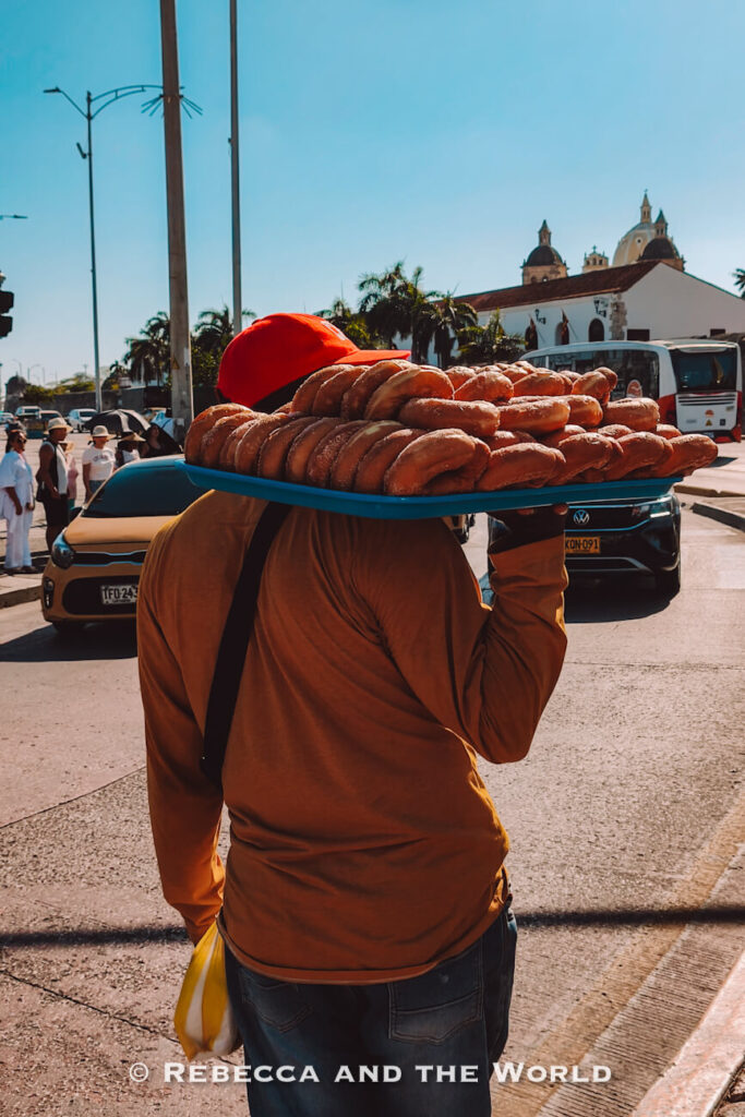A street vendor wearing a bright orange cap balances a large tray of donuts on one shoulder while walking along a busy road with cars and a white church in the background during a Cartagena street food tour.