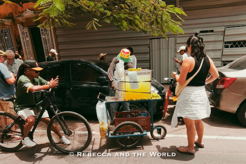 A street juice vendor stands beside a small cart filled with limes and plastic cups while customers gather on a shaded city street during a Cartagena food tour.