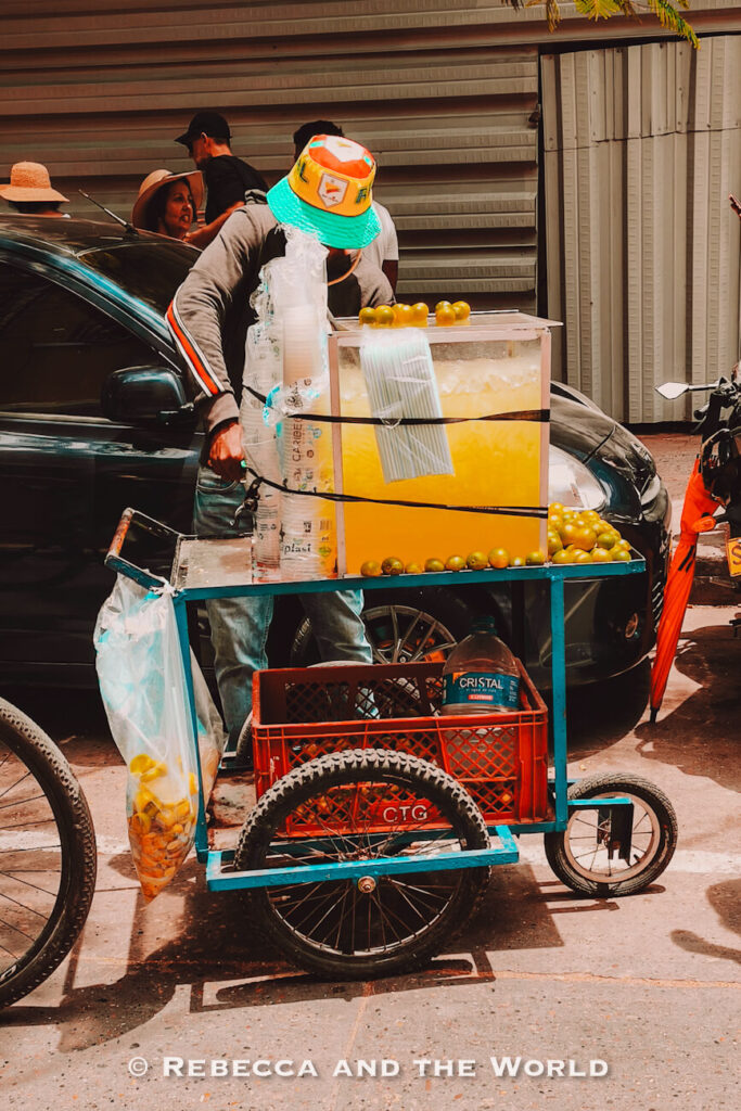 A close-up of a street drink cart loaded with large containers of lime juice, stacks of plastic cups, and fresh limes, parked beside a dark car in Cartagena.