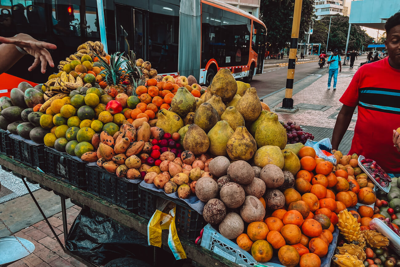 A large roadside fruit cart displays heaps of oranges, pears, bananas, coconuts, and tropical fruit beside a city bus stop during a Cartagena food tour.