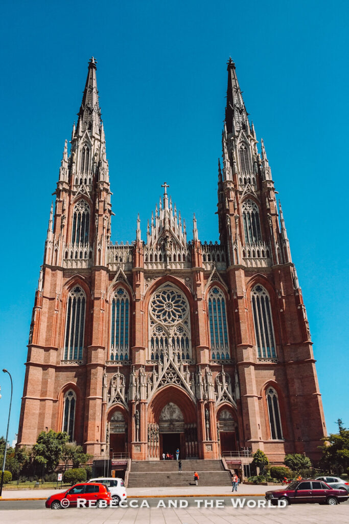 Front view of a large Gothic-style cathedral with two tall spires rising against a bright blue sky. Ornate stonework, arched windows, and a circular rose window decorate the façade. A wide staircase leads to the main entrance, where a few people are visible, while several cars are parked along the street in front. This impressive church is a popular sight on a day trip from Buenos Aires to La Plata.