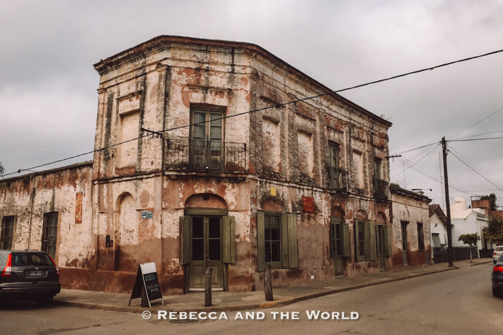 Corner view of a weathered two-story historic building in San Antonio de Areco, with peeling plaster walls, green wooden shutters, wrought-iron balconies, and several doorways opening onto a quiet street. Cars are parked along the curb, utility poles and wires run overhead, and the cloudy sky adds to the aged atmosphere. This colonial-era streetscape is a charming scene often encountered in San Antonio de Areco, one of the best day trips from Buenos Aires.