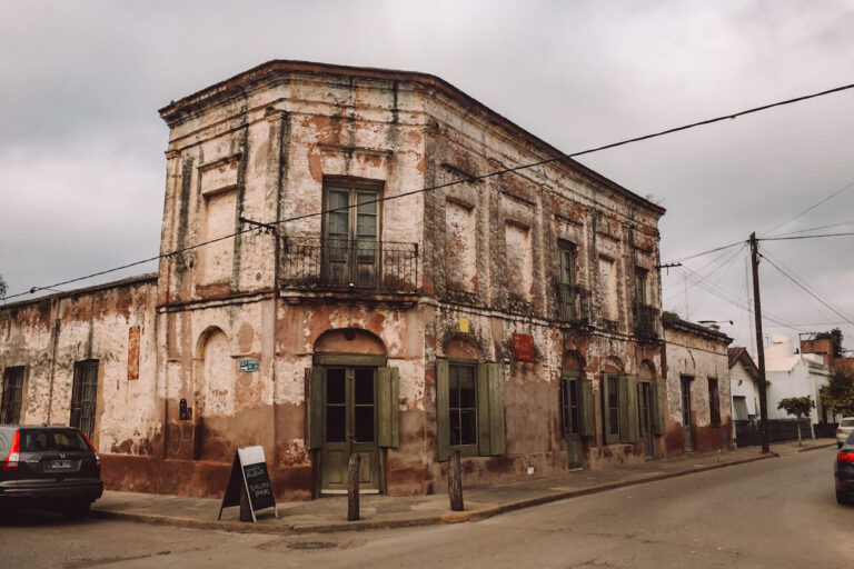 Corner view of a weathered two-story historic building in San Antonio de Areco, with peeling plaster walls, green wooden shutters, wrought-iron balconies, and several doorways opening onto a quiet street. Cars are parked along the curb, utility poles and wires run overhead, and the cloudy sky adds to the aged atmosphere. This colonial-era streetscape is a charming scene often encountered in San Antonio de Areco, one of the best day trips from Buenos Aires.