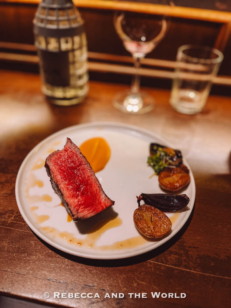 Medium-rare steak served on a white plate with roasted vegetables and sauce at Fogon Asado in Buenos Aires, with wine glasses and a bottle blurred in the background.