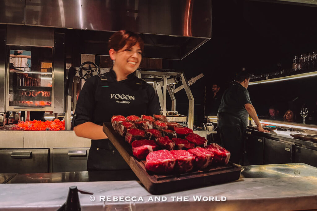 Chef carrying a large tray of raw steaks through the open kitchen at Fogon Asado restaurant in Buenos Aires.