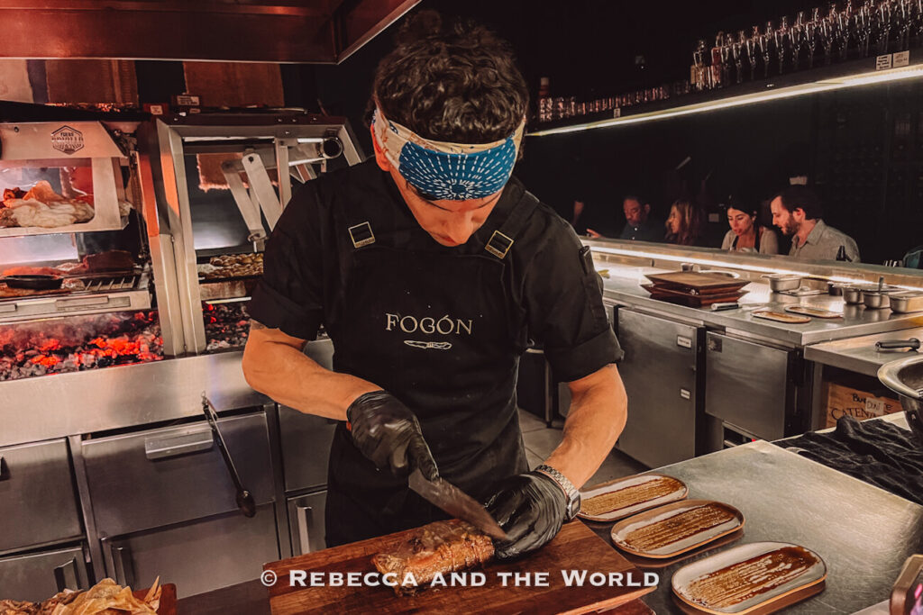Chef slicing grilled meat at an open-fire kitchen counter inside Fogon Asado, part of a memorable Buenos Aires itinerary for food lovers.