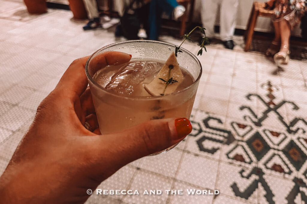 Hand holding a cocktail with ice and a citrus garnish inside Fogon Asado restaurant in Buenos Aires, with patterned tile flooring and seated guests in the background.