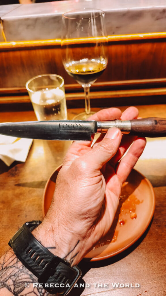 Hand (the author's husband's) holding a Fogon-branded steak knife above a wooden dining counter with a wine glass and water glass in the background.