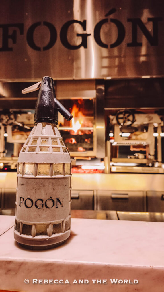 Fogon-branded siphon bottle displayed in front of the open-fire grill inside Fogon Asado restaurant.