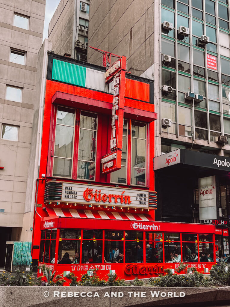 A bright red corner building with large windows and vintage-style signage stands between tall gray office buildings on a busy city street in Buenos Aires. The restaurant façade reads “Guerrin” in glowing red letters, with striped red-and-white awnings and people dining inside at street level. To the right, a modern black entrance sign for Teatro Apolo contrasts with the colorful historic storefront. The urban scene captures the lively atmosphere often featured on the best food tours in Buenos Aires.