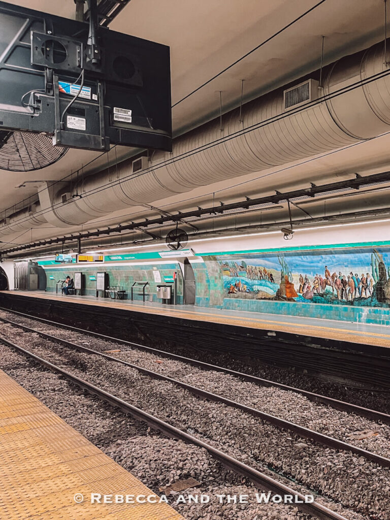 An underground subway platform in Buenos Aires features train tracks, turquoise station walls, and a long mural showing historical scenes. Overhead pipes and lighting run across the ceiling, showing a typical station used for public transport in Buenos Aires.