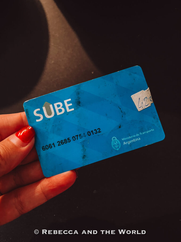 A hand with red-painted nails holds a blue SUBE transport card against a dark background. The card is used to pay for buses, trains, and the subway system, making it an essential part of how to get around Buenos Aires using public transport.