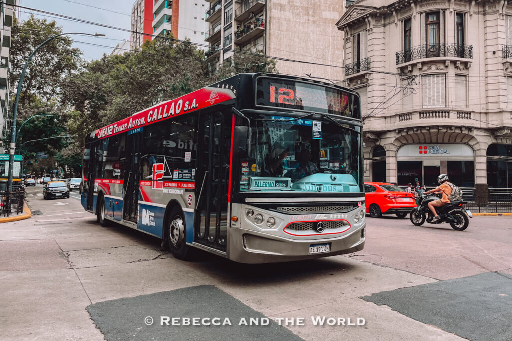 A red, blue, and silver city bus marked with route number 12 turns through an intersection lined with apartment buildings and trees in Buenos Aires. Cars and a motorbike share the road, capturing a busy scene of everyday Buenos Aires transport.