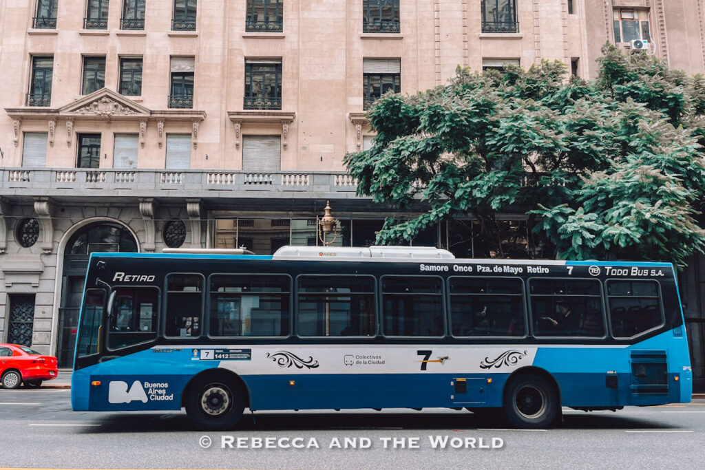 A blue city bus drives along a wide street in Buenos Aires, passing historic stone buildings and large green trees. The bus displays route number 7 and “Retiro” on the front side panel, illustrating public transport in Buenos Aires and giving a realistic look at how to get around Buenos Aires by local bus.