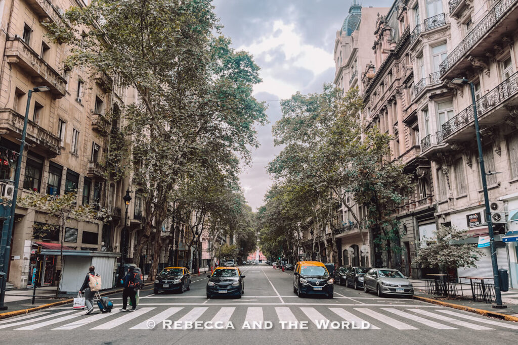 Wide view of a tree-lined boulevard in Buenos Aires, with historic apartment buildings and ornate balconies on both sides of the street under a cloudy sky. Several black-and-yellow taxis and parked cars line the road, while two pedestrians cross at a zebra crossing in the foreground carrying luggage and shopping bags. Taxis are one of the easiest ways to get around Buenos Aires.
