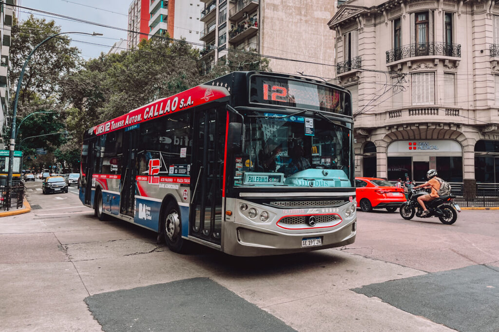 A red, blue, and silver city bus marked with route number 12 turns through an intersection lined with apartment buildings and trees in Buenos Aires. Cars and a motorbike share the road, capturing a busy scene of everyday Buenos Aires transport.