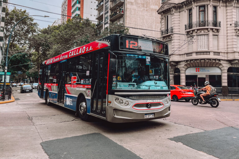 A red, blue, and silver city bus marked with route number 12 turns through an intersection lined with apartment buildings and trees in Buenos Aires. Cars and a motorbike share the road, capturing a busy scene of everyday Buenos Aires transport.