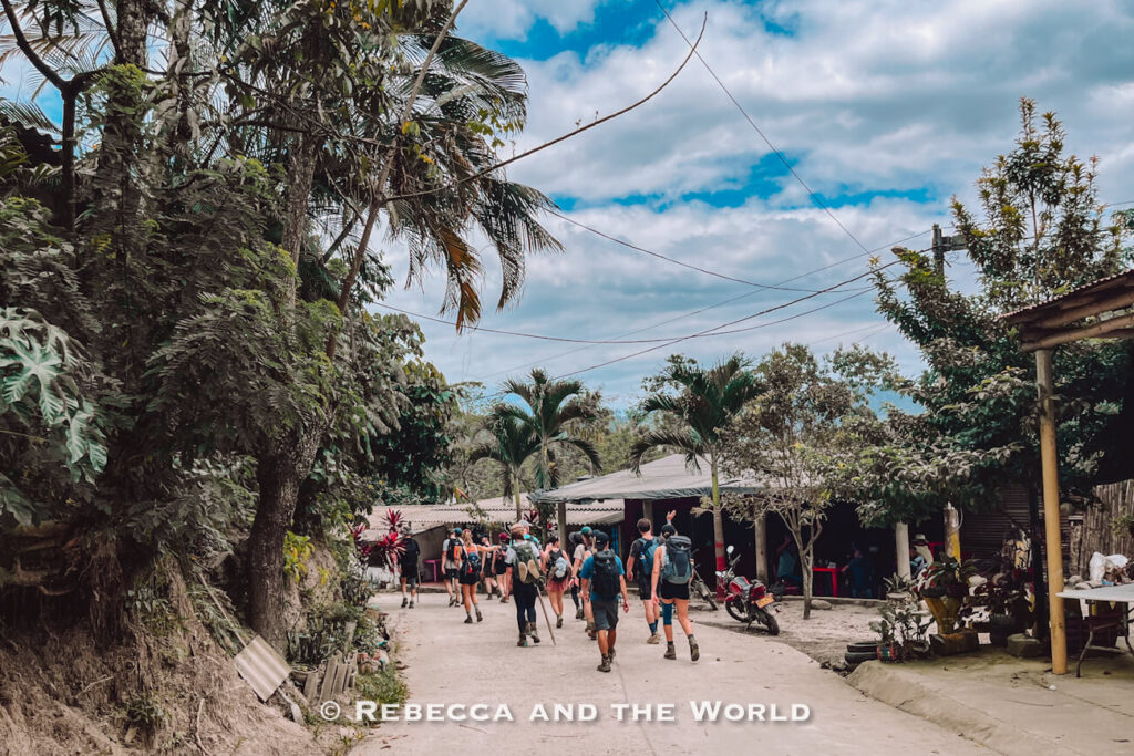 Group of hikers walking through a small jungle village at the start of the Lost City trek in Colombia, surrounded by palm trees, rustic homes, and cloudy skies during the Ciudad Perdida hike.