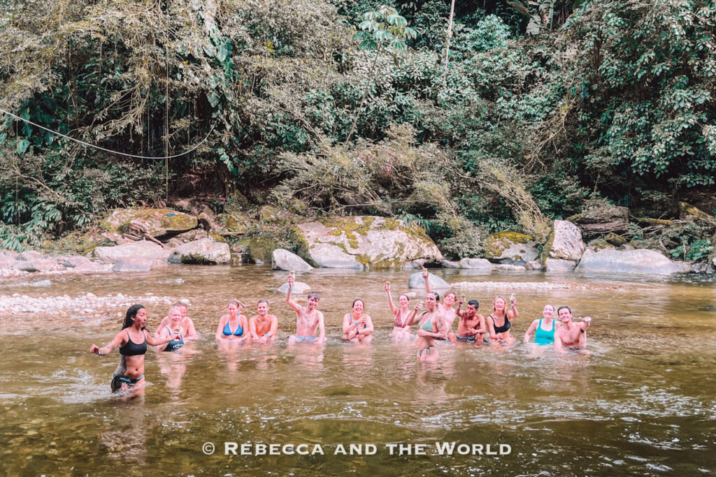 Travelers cooling off in a shallow jungle river during the Lost City trek, with dense rainforest and large rocks surrounding the swimming spot on the Ciudad Perdida hike.