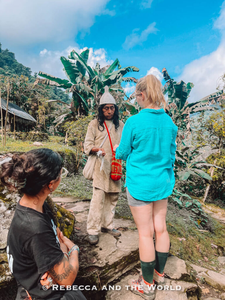 Traveler speaking with an Indigenous Kogi person in a tropical village during the Ciudad Perdida hike, with banana trees and mountain scenery in the background on the Lost City trek.