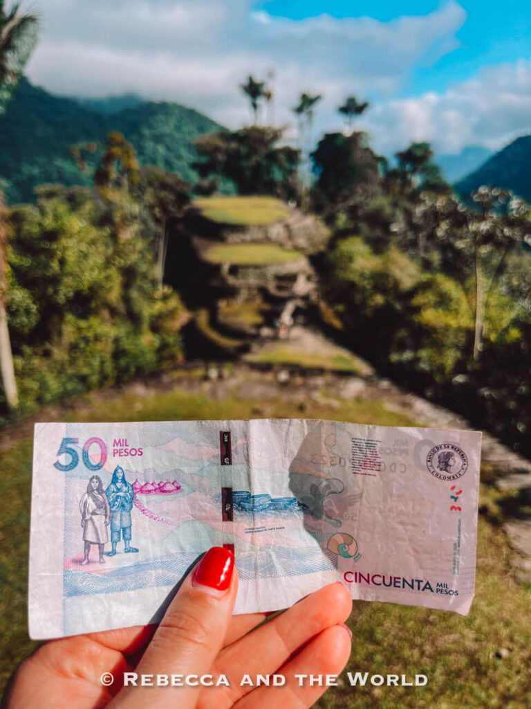 Hand holding a 50,000 Colombian peso note with the ancient terraces of Ciudad Perdida blurred in the background during the Lost City hike.