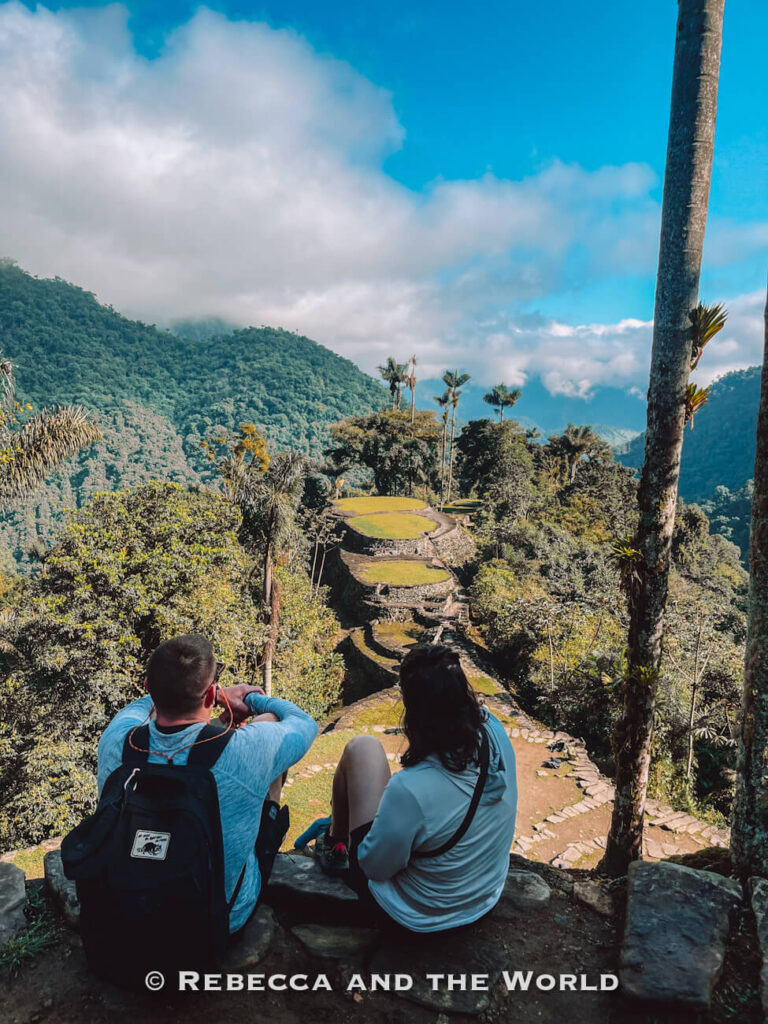 Two hikers (the author and her husband) sitting on a stone ledge overlooking the terraced ruins of Ciudad Perdida surrounded by rainforest during the Lost City trek.