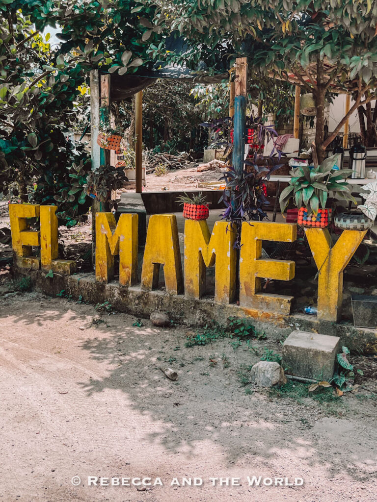 Large yellow “El Mamey” sign beside a rural road surrounded by trees, marking the starting point of the Lost City trek.
