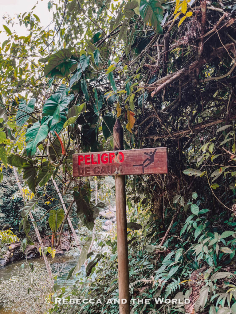 Wooden warning sign reading “Peligro de Caída” beside a jungle river trail during the Ciudad Perdida trek in Colombia.