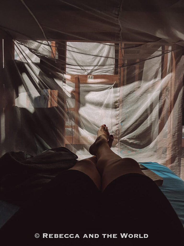 A view from inside a dimly lit sleeping area shows a person lying on a bed beneath hanging mosquito nets, with sunlight filtering through the fabric walls.