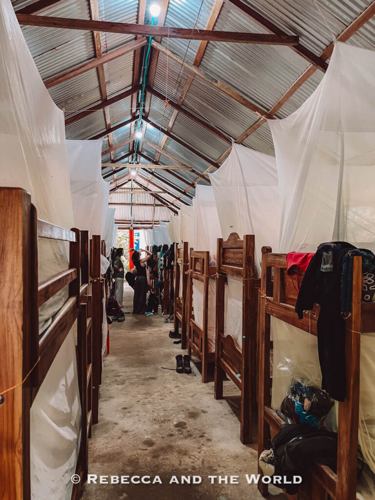 A long dormitory-style room contains rows of wooden bunk beds covered with white mosquito nets, with backpacks and clothing hanging nearby. This is the accommodation on the Ciudad Perdida hike.