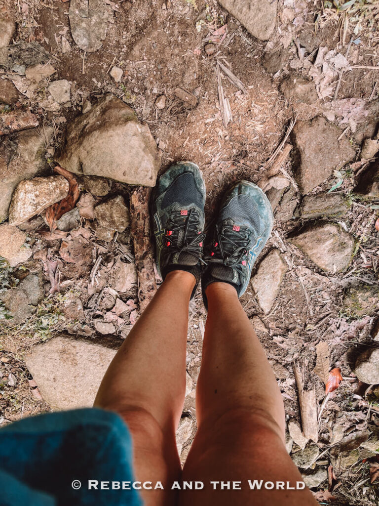 A person (the author of this article) wearing trail shoes stands on a dirt and rock-covered hiking path, photographed from above. One of the decisions you'll need to make for the Lost City trek packing list is where you wear trail runners or hiking boots.