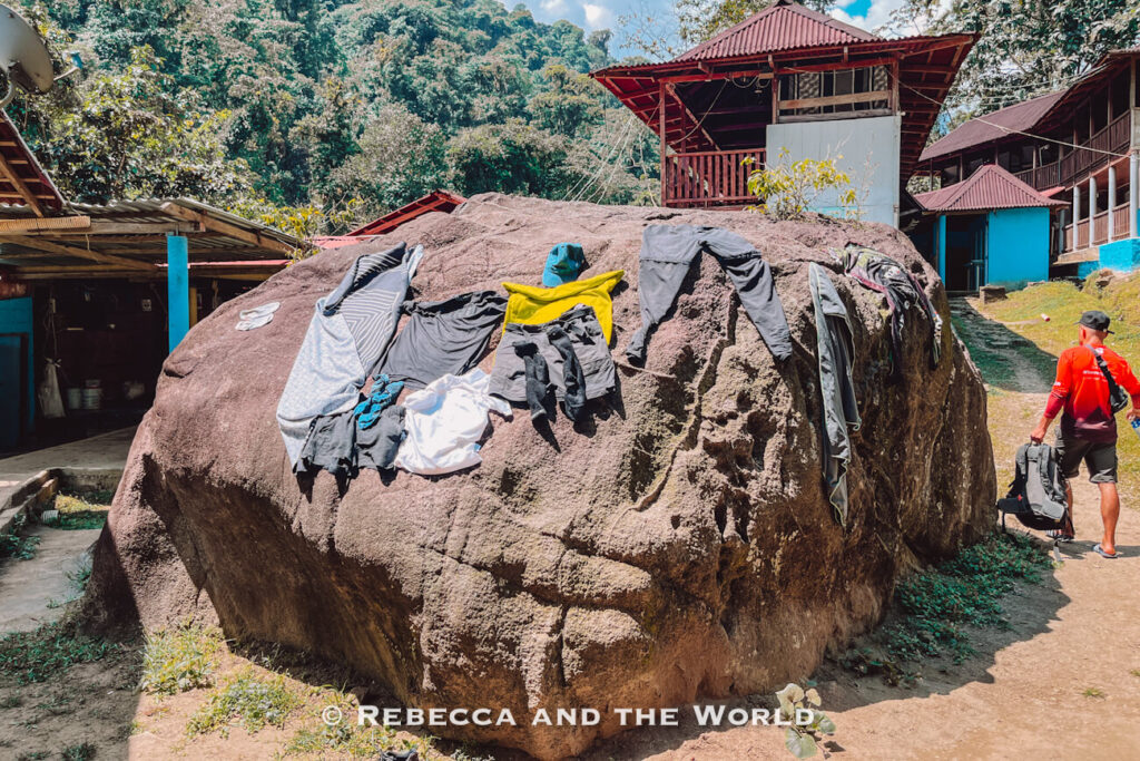 Clothes and gear are spread out to dry on top of a large boulder in a small mountain village surrounded by rustic wooden buildings. The Lost City trek is humid so clothes will rarely dry!