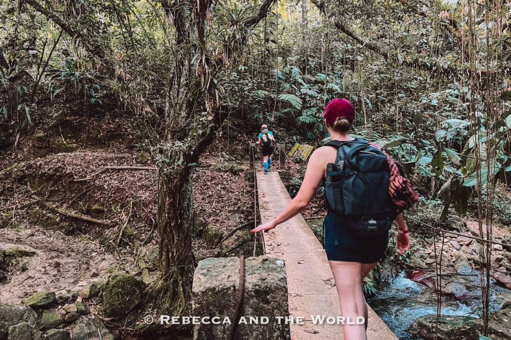 Two hikers wearing backpacks cross a narrow concrete footbridge over a stream in dense rainforest. There are several bridges and river crossings along the Lost City trek.
