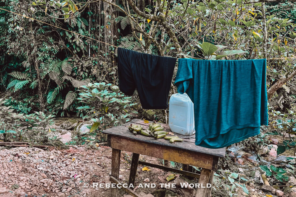 Clothes hang on a line above a wooden table holding bananas and a water container beside a rocky stream in the jungle.