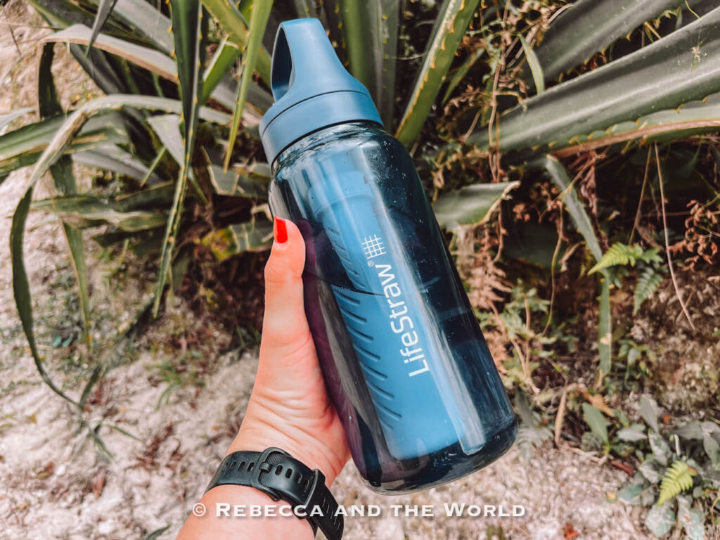 A person holds a blue reusable water bottle outdoors near tropical plants and rocks. The LifeStraw bottle is a great addition to a Lost City trek packing list.