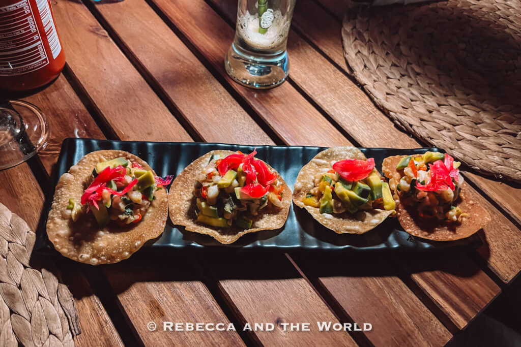 Four crispy tostadas are arranged on a black rectangular platter atop a wooden slatted table. Each tostada is topped with diced seafood, avocado chunks, pickled red onions, and vegetables, creating a colorful presentation. Glassware and woven placemats are partially visible around the table, suggesting an outdoor restaurant setting in Palomino Colombia.