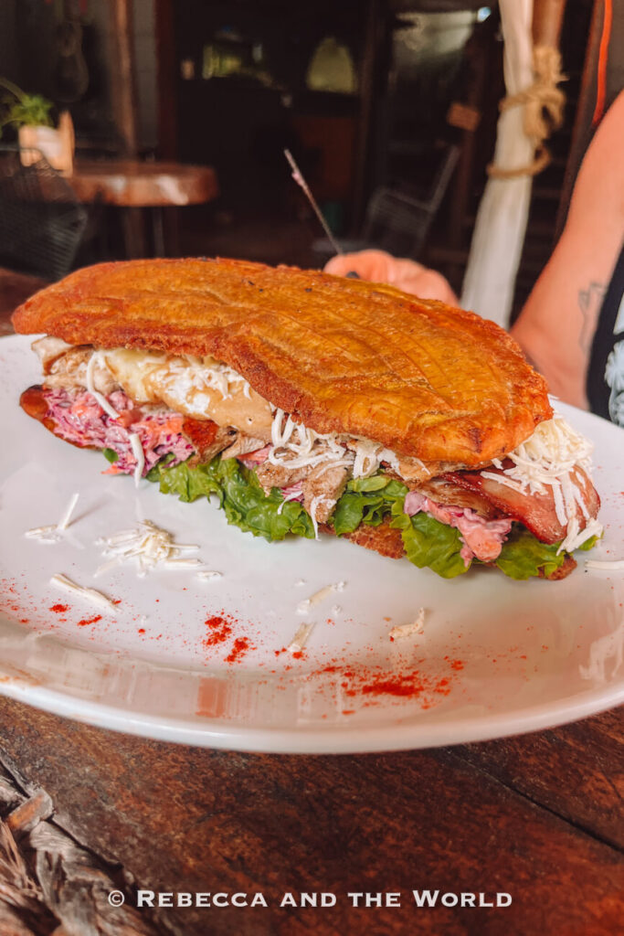 A large toasted patacone "burger" filled with grilled meat, lettuce, shredded cheese, bacon, and a creamy slaw sits on a white plate dusted with red seasoning. A person holding the plate is partially visible in the background. This hearty meal is one of the many food experiences featured in a Palomino travel guide.