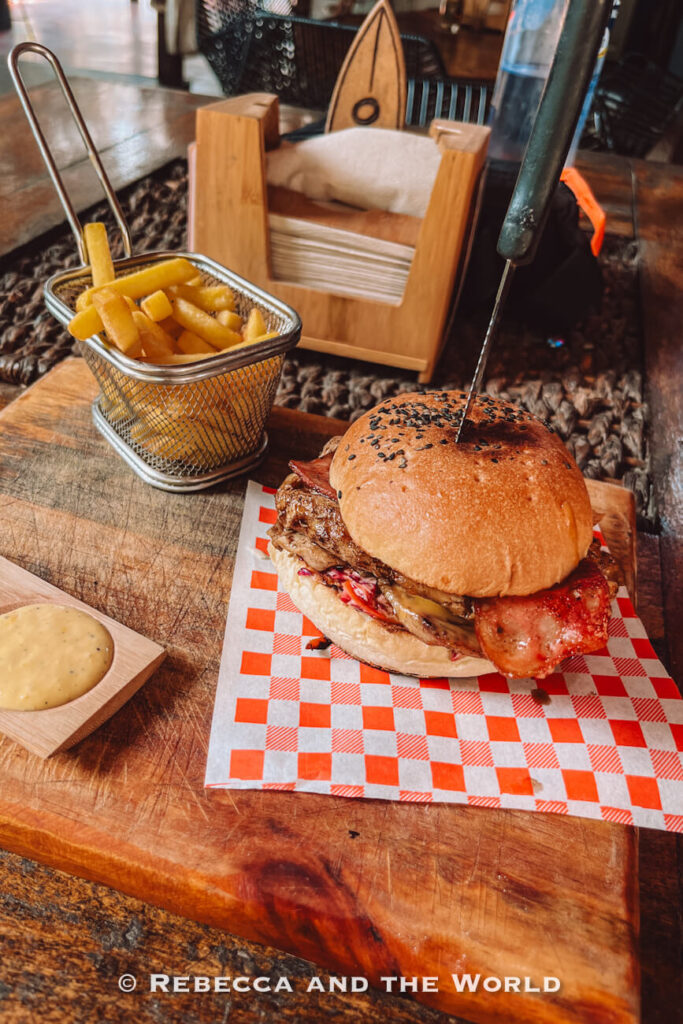 A chicken burger topped with bacon and grilled meat is served on checkered paper beside a metal basket of thick-cut fries. A small dish of dipping sauce sits on the wooden table, while the rustic restaurant interior fades into the background. Sampling local restaurants is among the popular things to do in Palomino.