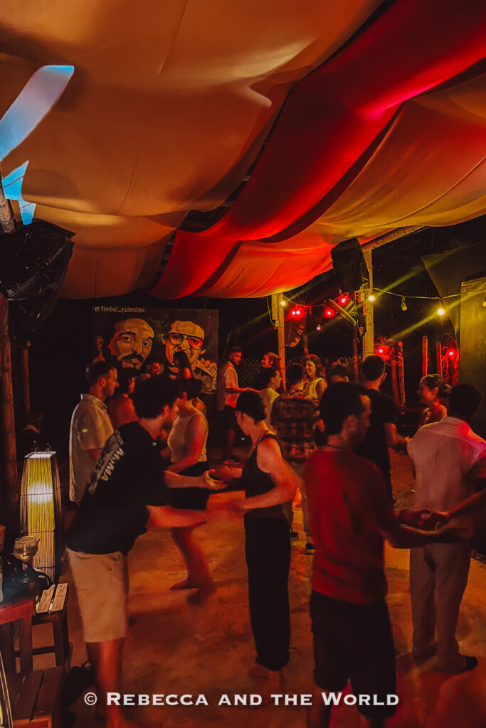 Several people salsa dancing under flowing fabric draped across the ceiling in a dimly lit venue illuminated by red and warm-colored lights. A mural decorates the back wall, and the lively atmosphere captures Palomino’s nightlife scene. Experiencing local music and dancing is one answer to what to do in Palomino after sunset.