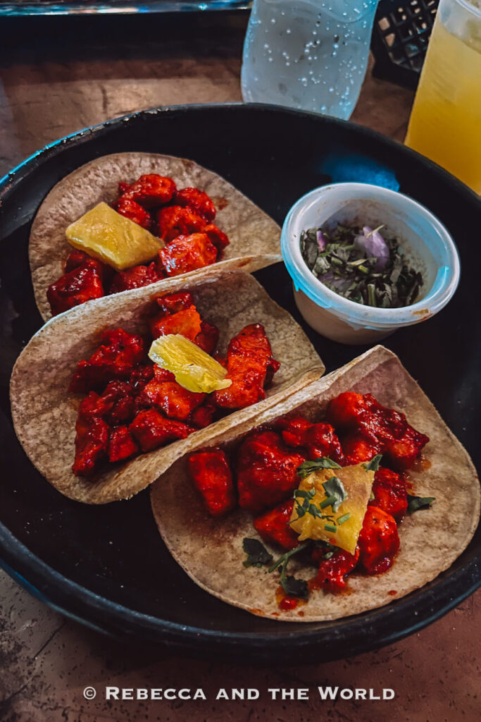 Three soft-shell tacos filled with bright red grilled meat are served on a black plate. Each taco is garnished with pineapple, while a small container of chopped onions and herbs sits alongside them. Food lovers often include restaurants like this in a Palomino itinerary.