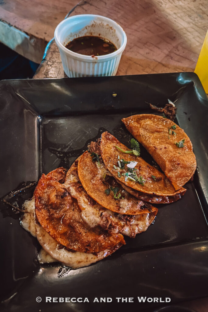 Four folded tacos stuffed with shredded meat and melted cheese are arranged on a black plate. A small cup of dark dipping broth sits behind them, and melted cheese spills from the edges of the tortillas. This popular dish can be found at eateries throughout Palomino Colombia.