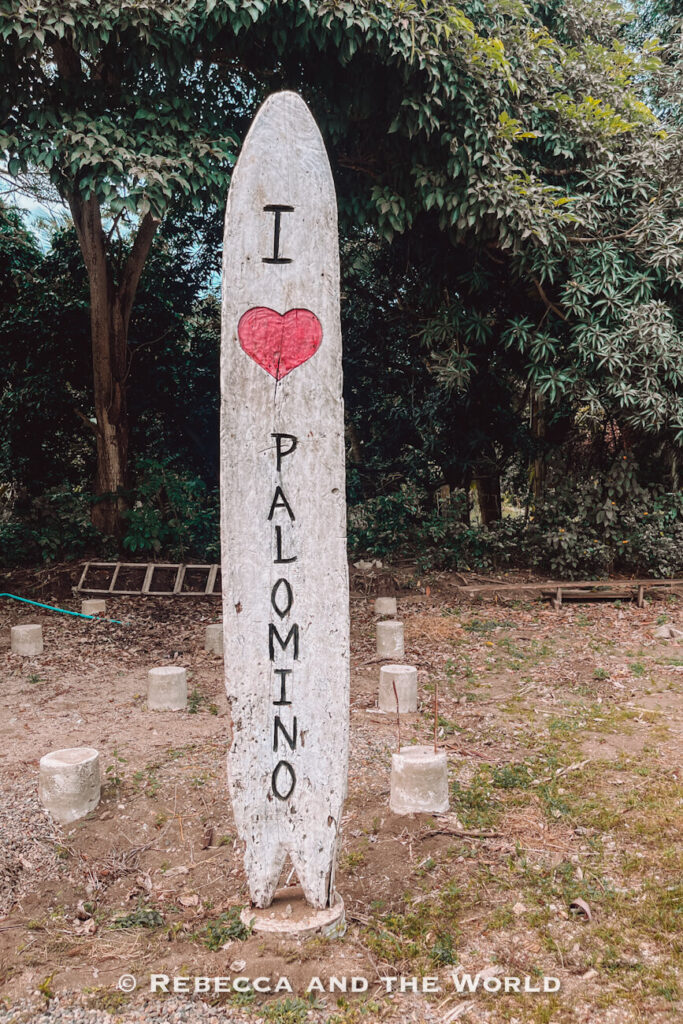 A weathered surfboard-shaped sign stands upright among trees and greenery. Painted on the board are the words “I ♥ Palomino,” with a red heart replacing the word “love.” Taking photos at landmarks like this is a popular choice when deciding what to do in Palomino.