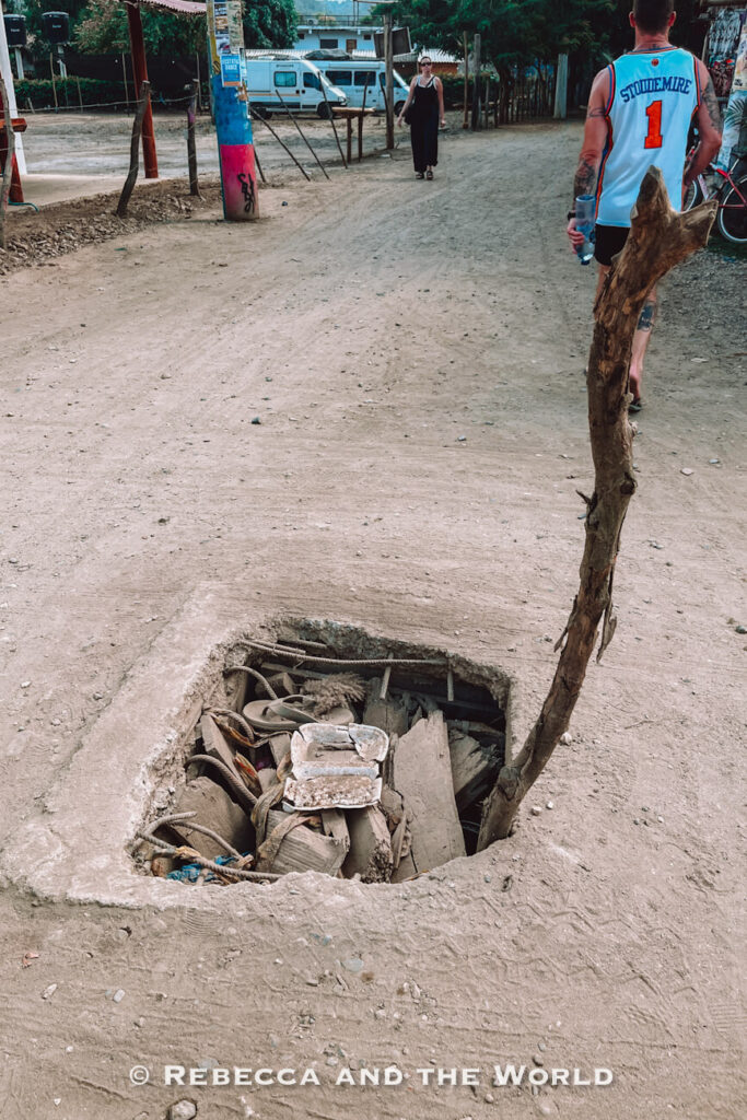 An uncovered hole filled with debris and construction materials sits in the middle of a dusty road. A stick has been placed upright beside it as a warning marker, while pedestrians walk nearby. This image shows the rustic infrastructure that visitors may encounter during a Palomino itinerary.