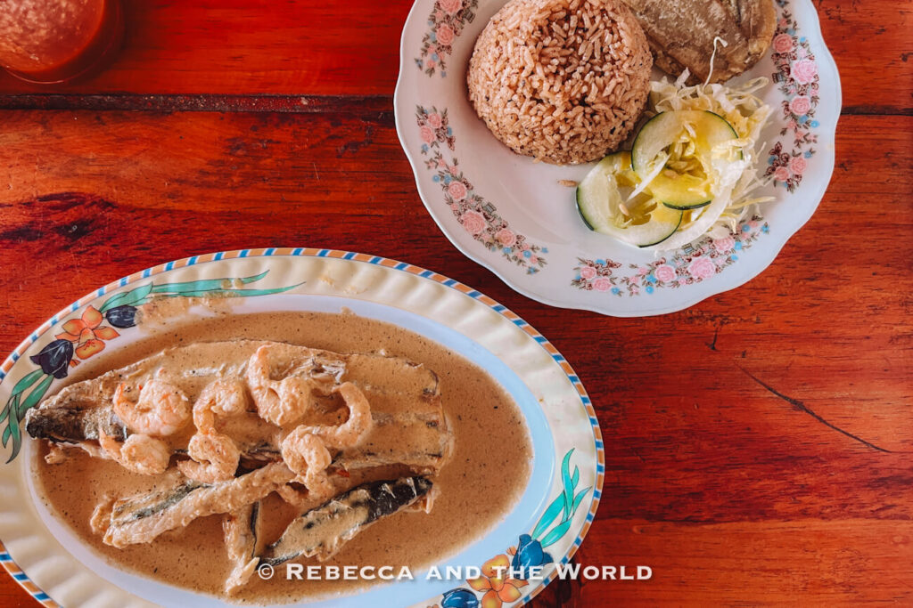 A whole fish covered in a creamy sauce and topped with shrimp is served on a decorative plate. A side plate holds rice, salad, and fried plantain, all resting on a bright red wooden table. Traditional Caribbean cuisine is often featured in a Palomino travel guide.