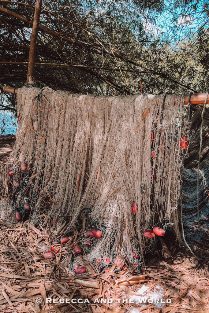 Large fishing nets hang from a wooden pole beneath dense coastal vegetation, with red floats attached throughout the tangled mesh. Dry leaves cover the ground below, highlighting the area's connection to local fishing traditions in Palomino Colombia.