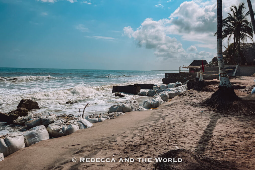Sandbags and concrete barriers line a section of beach where waves crash against the shoreline. Palm trees and beachfront buildings stand nearby, illustrating efforts to protect coastal areas in Palomino Colombia.