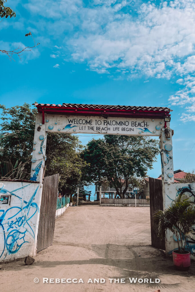 A large white gateway painted with blue wave designs welcomes visitors to the beach. The sign reads “Welcome to Palomino Beach – Let the Beach Life Begin,” with sandy pathways leading toward the ocean beyond. Landmarks like this often appear in a Palomino travel guide.