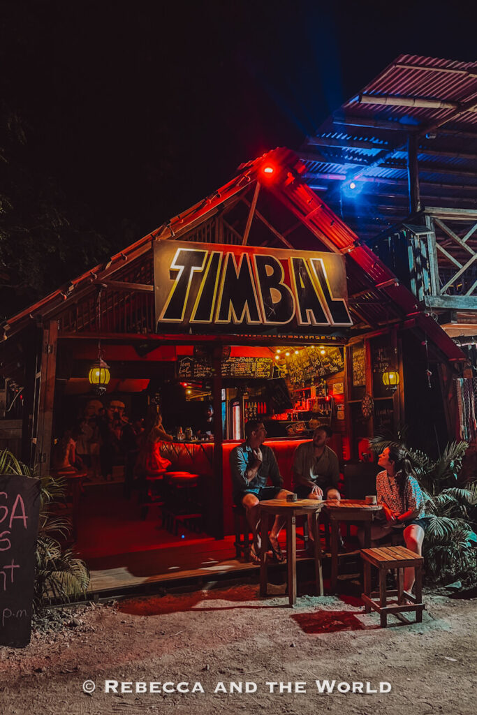 A small open-air bar glows with red and blue lighting after dark. Several people sit at outdoor tables while others gather inside beneath a large sign reading “TIMBAL.” Enjoying nightlife spots like this is one of the popular things to do in Palomino.
