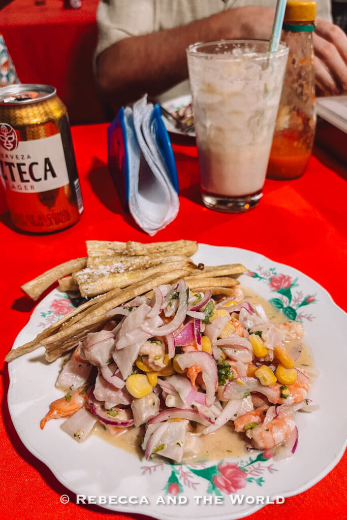 A plate of ceviche made with fish, shrimp, red onion, corn, and herbs sits on a bright red tablecloth alongside fried plantain strips. A beer, hot sauce, and a cold drink complete the meal. Sampling fresh seafood is a delicious option when considering what to do in Palomino.
