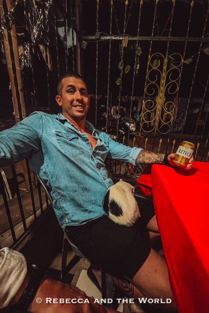 A man - the author's husband - sits at a table holding a can of beer while a black-and-white cat curls up on his lap. The cozy, dimly lit restaurant setting creates a relaxed atmosphere that reflects the laid-back pace of a Palomino itinerary.